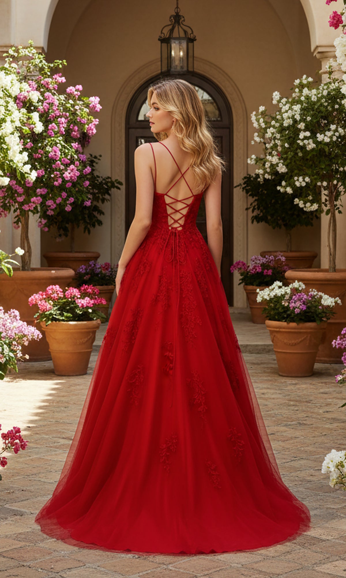 A woman wearing the PromGirl Exclusive 250228 long red prom dress with lace embroidery and an adjustable lace-up back stands among potted flowering plants, facing away from the camera.